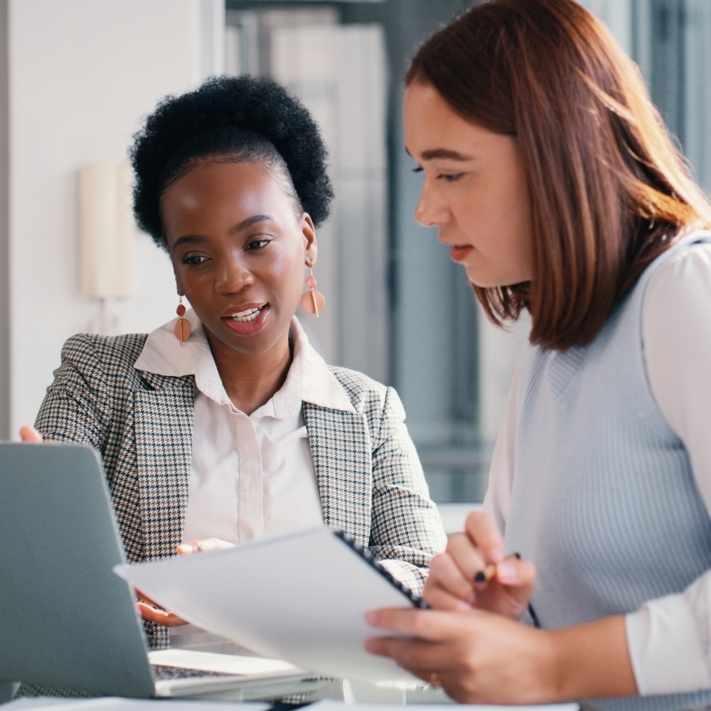 One person with long red hair and another with short dark hair seated at desk looking at open laptop computer