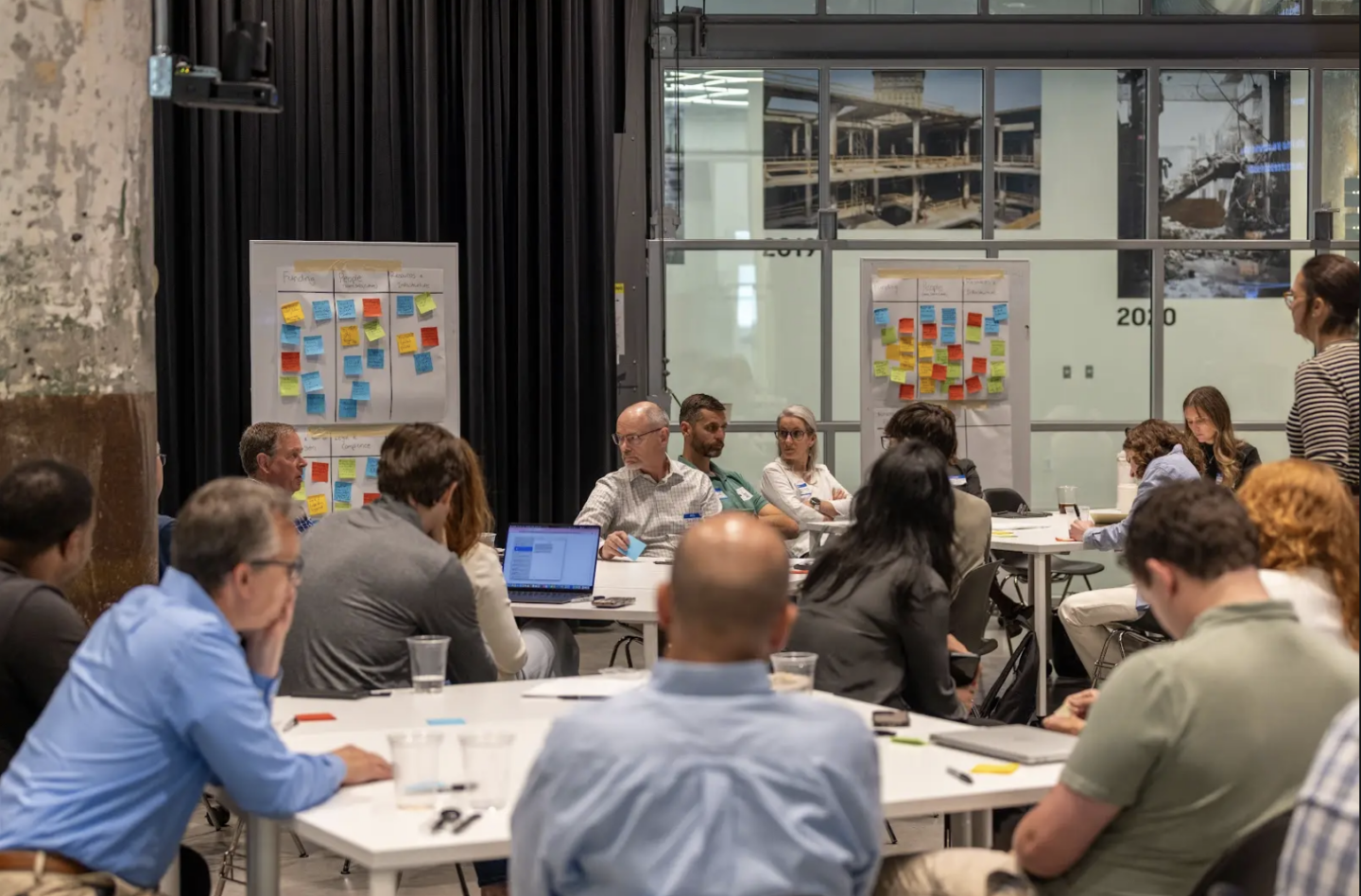 People seated around tables with laptop computers and poster boards with multi-colored sticky notes in background