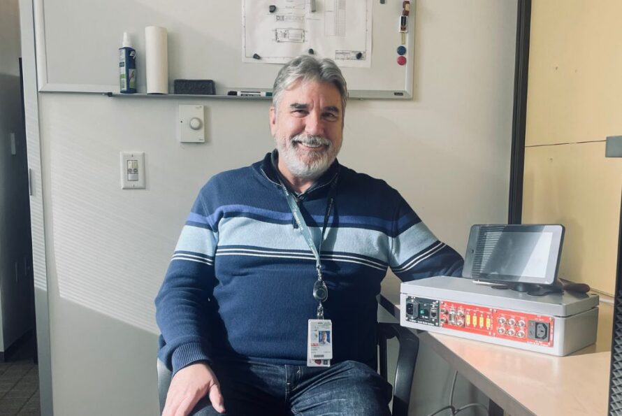 Person with short gray hair and beard wearing a blue sweater sitting at desk smiling