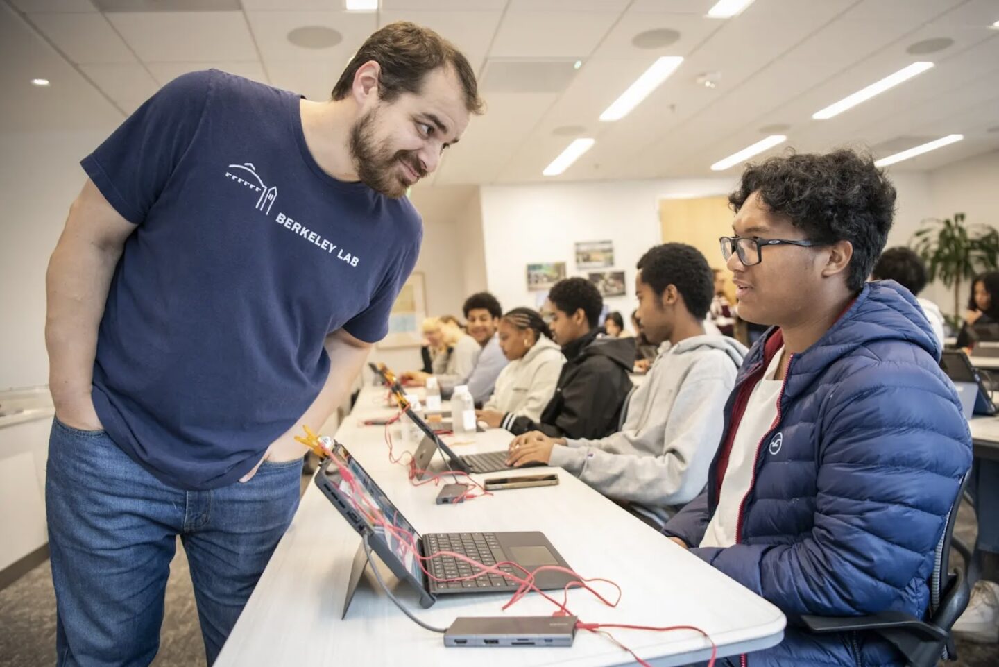 Person with dark hair and beard leaning over another person's laptop that is sitting on a table