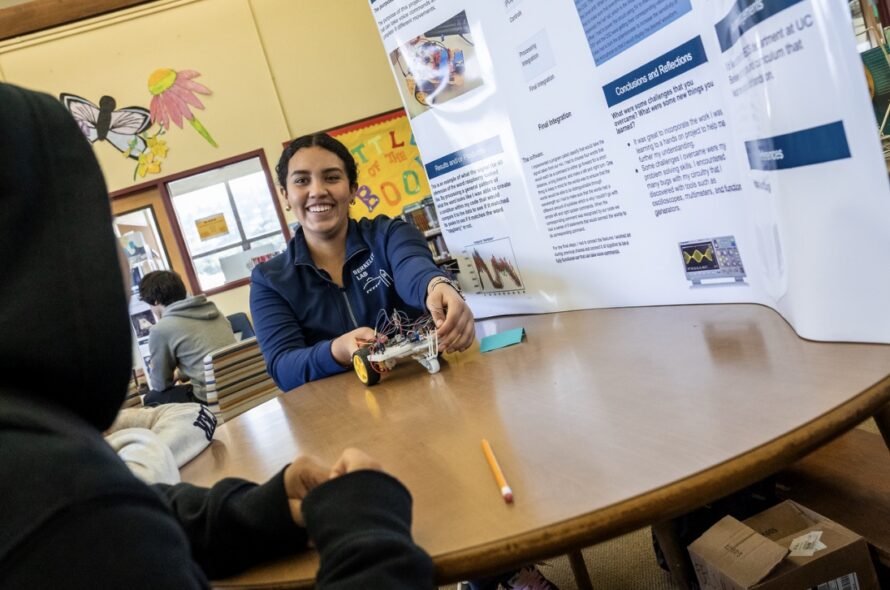 Person with black hair wearing a blue shirt seated at round wooden table adjacent to poster boards