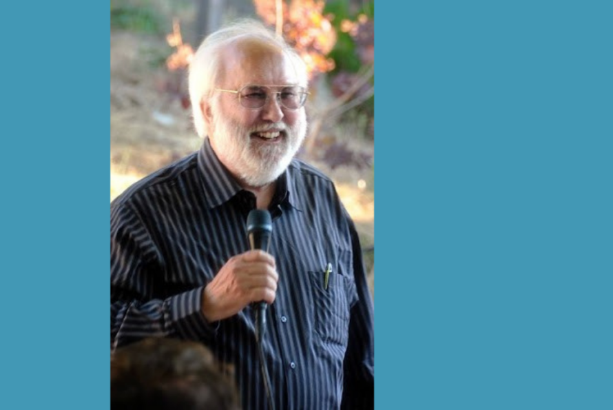 Photo of smiling person with gray hair and beard wearing a dark blue dress shirt and holding a microphone in one hand