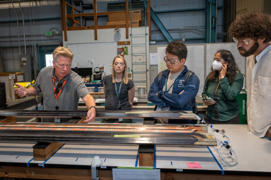 Five persons wearing protective eyewear and standing over a lab table while one person makes adjustments to instrument
