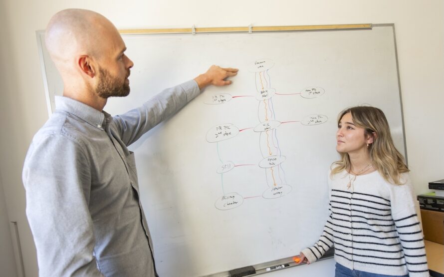 Two persons standing in front of white board with one person pointing at the board