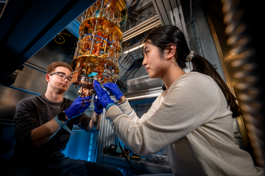One person with long black hair wearing tan sweater and protective gloves and another person with red hair and black glasses wearing black sweater adjusting cylindrical scientific instrument suspended from above