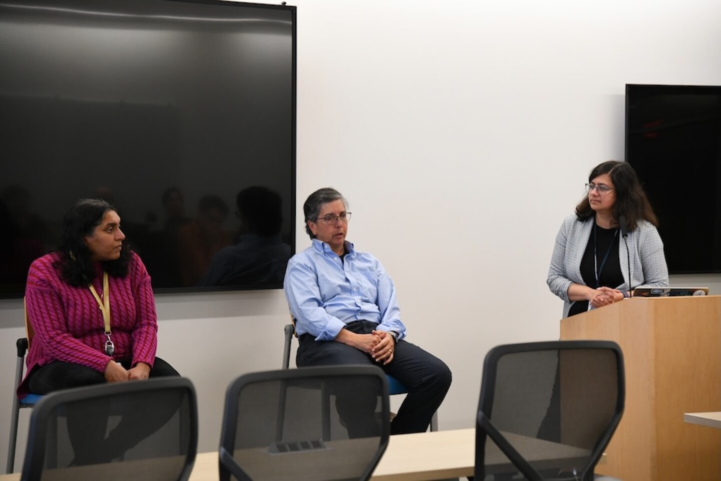 Two people sitting in chairs next to a podium listening to a third person standing and facing the two other persons