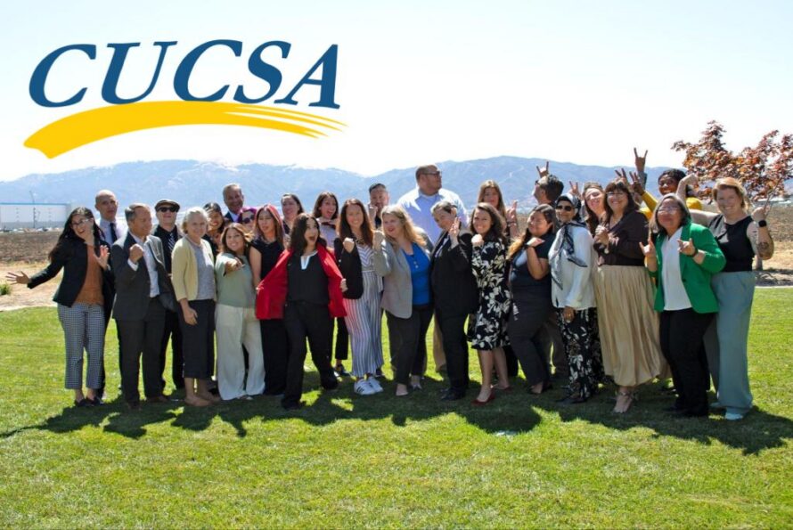 Group of people standing together on grass with hills and sky in background and a "CUCSA" logo in upper-left.
