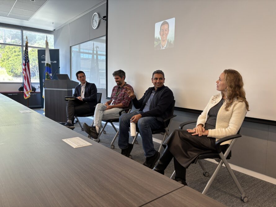 Four persons sitting at large conference room table with image of person projected onto wall behind them