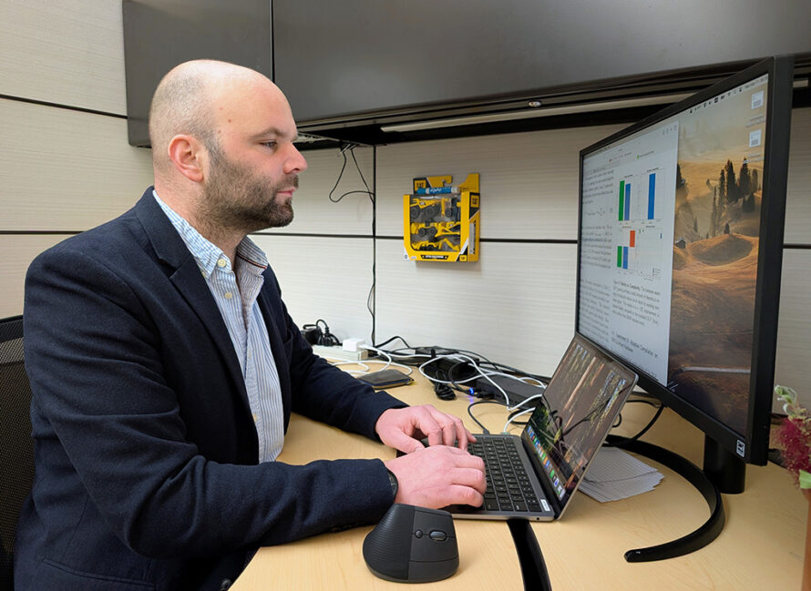 Person with black beard wearing dark blue suit sitting in front of computer monitor looking at monitor while typing on keyboard