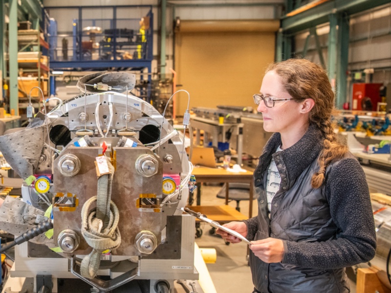A person wearing glasses and a braided ponytail stands in a workshop, holding a tablet while examining a large, complex metal machine with gauges, bolts, and attached cables. The industrial lab space behind her includes workbenches, tools, and equipment.