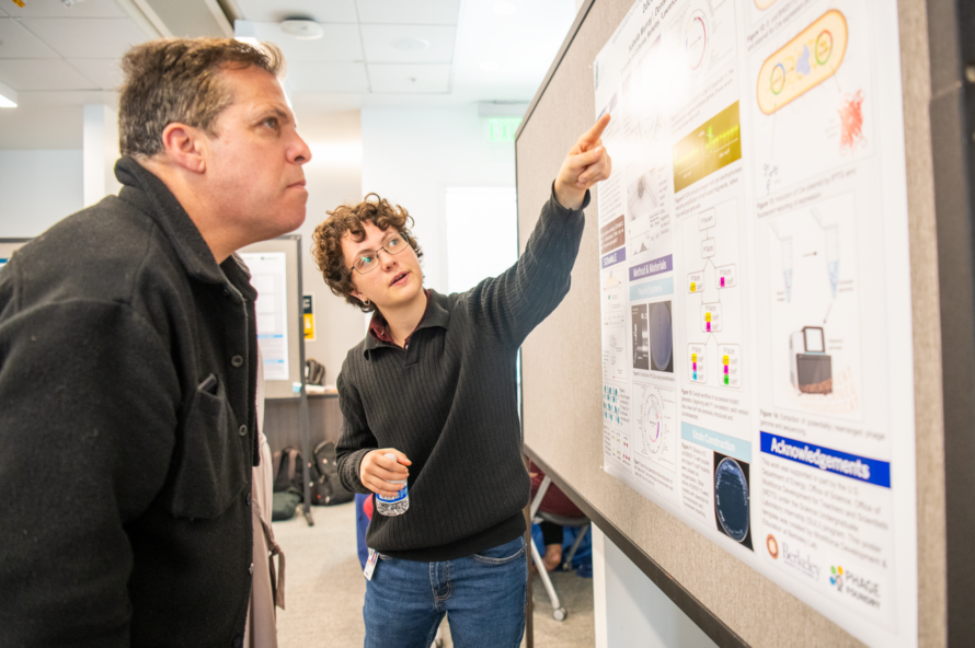 One person with short brown hair pointing to a poster board while second person with dark and gray hair observes