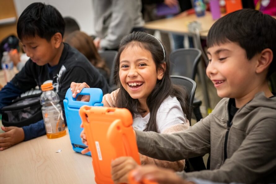 Two young person seated at table smiling, each of whom is holding up an orange and blue electronic device