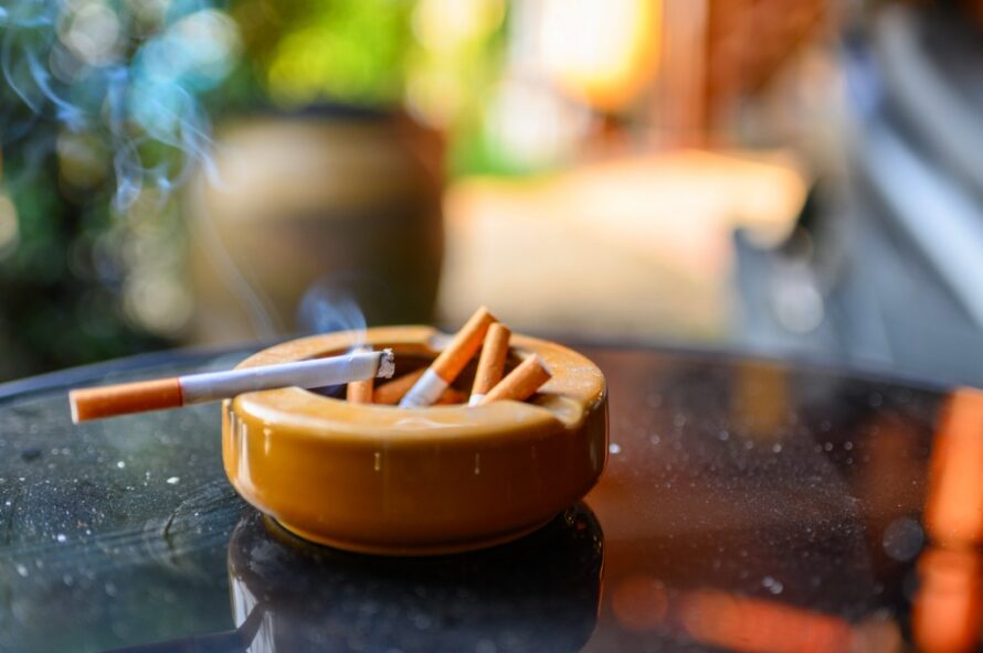 Brown ashtray sits atop a black table with several used cigarette butts inside and a lit cigarette resting on the side with smoking rising from it