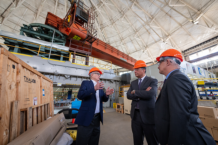 Three people wearing dark suit jackets and orange hard hats and safety glasses with large orange machinery above them.