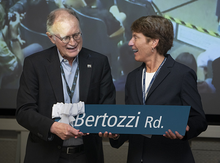 Two persons both wearing dark jackets holding a street sign in teal with white lettering.