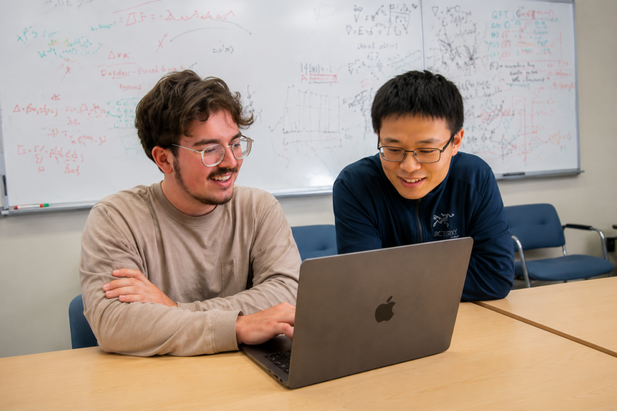 Two young persons seated in front of open laptop computer with white board in the background