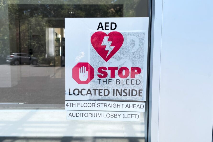 Sign on window that reads AED, Stop the Bleed Located Inside. 4th Floor Straight Ahead Auditorium Lobby (Left).