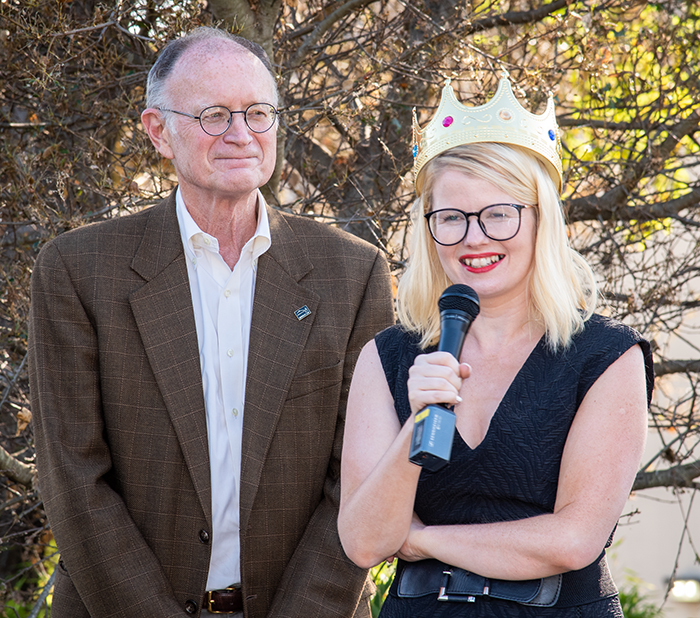 Two persons standing outside, one wearing dark suit jacket and looking off in distance while second person holding microphone wears a paper crown.
