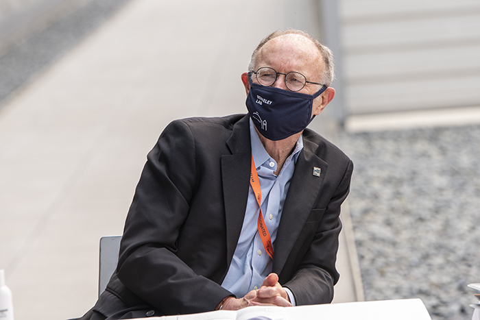 Person sitting at table wearing dark suit jacket, glasses, and face covering.
