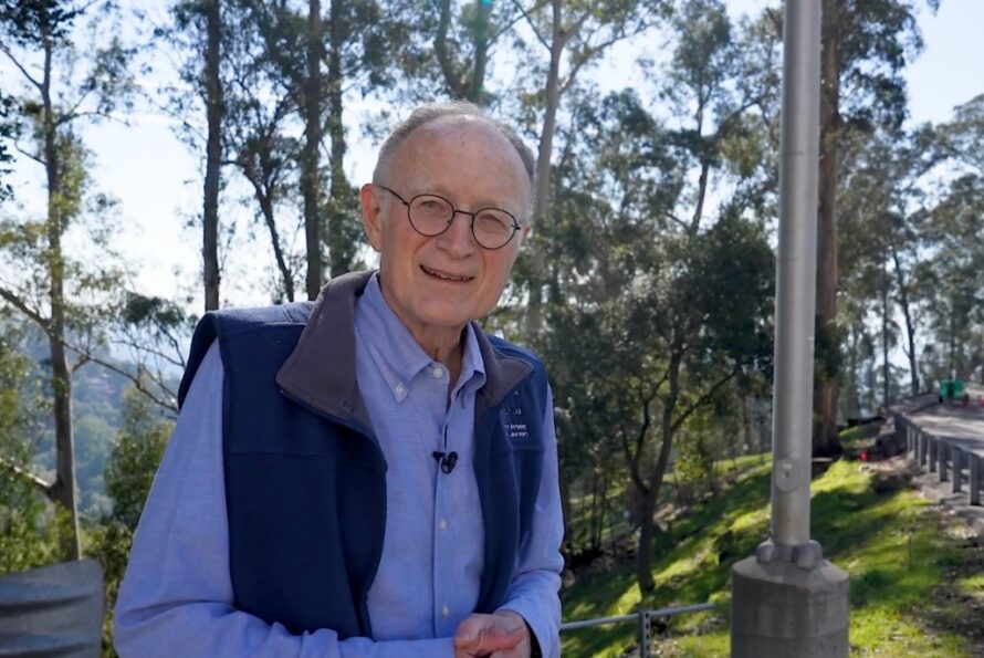 Mike Witherell in a blue-collared shirt and vest, smiling outdoors, with trees in the background.