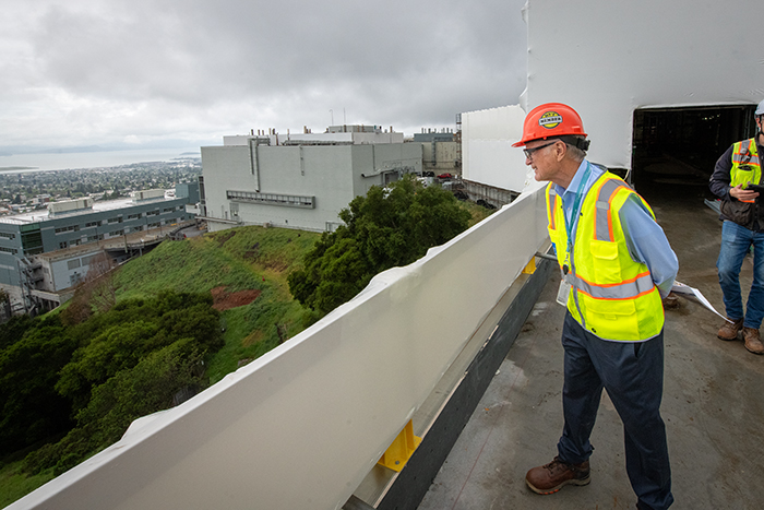 Person wearing safety vest, hard hat, and safety glasses standing on outside deck under construction looking over the edge.