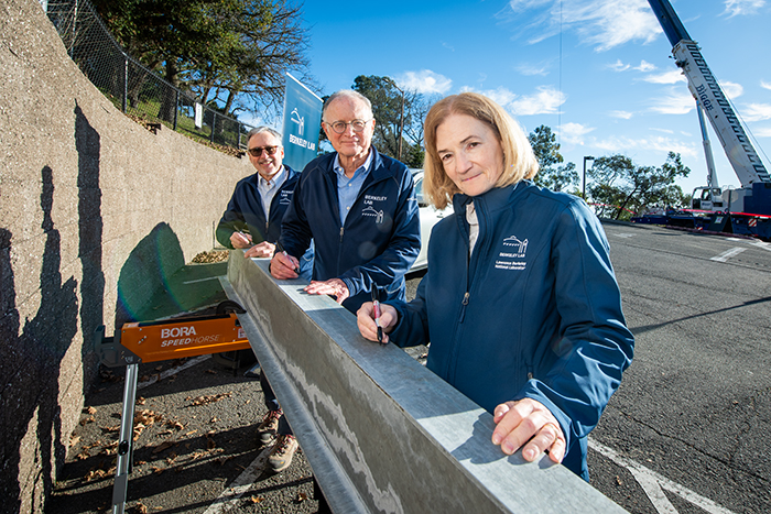 Three persons in a row each wearing dark blue jacket signing construction beam.