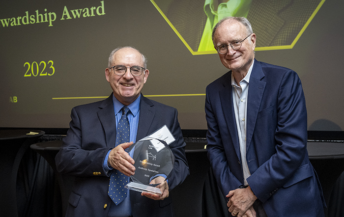 Two persons in dark blue suits standing on stage with one holding award.