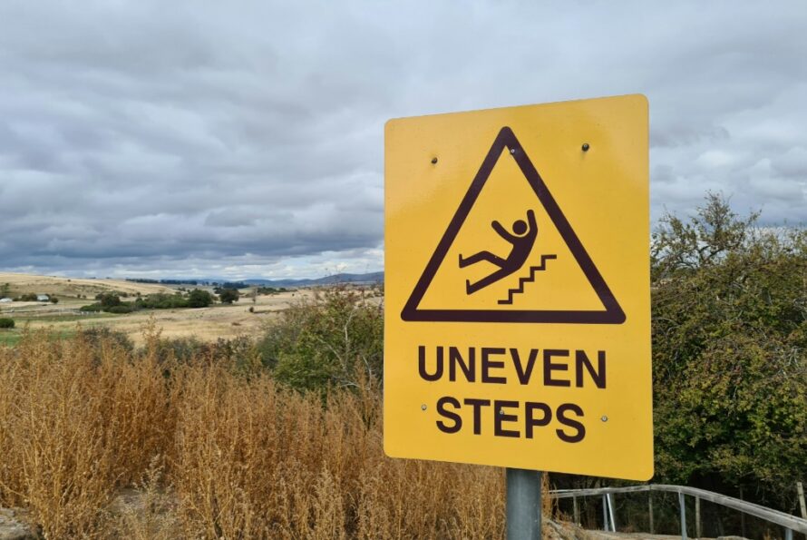 Yellow hazard sign displaying black triangle with person falling down stairs with words underneath "Uneven Steps"