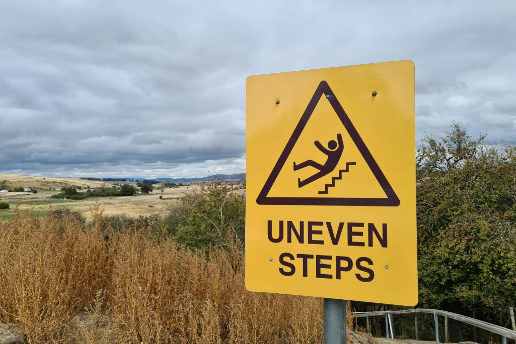 Yellow hazard sign displaying black triangle with person falling down stairs with words underneath "Uneven Steps"