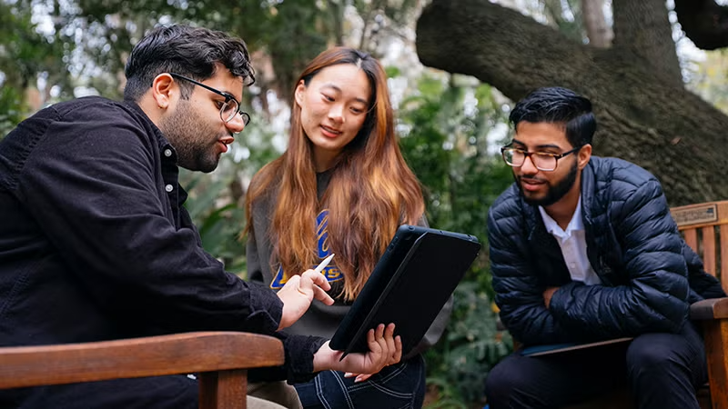 Three persons sitting outside on a bench all looking at a laptop computer the middle person is holding