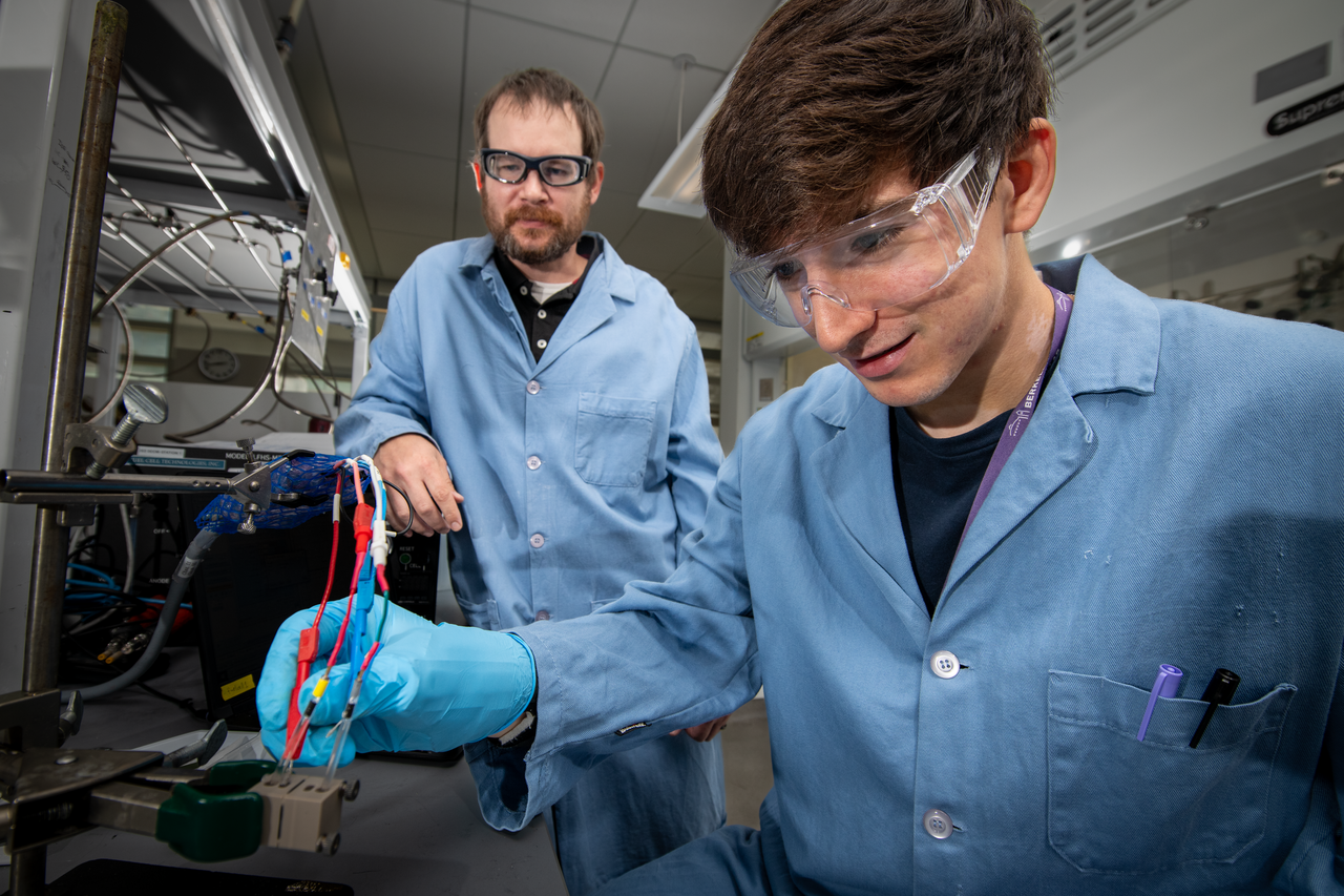 Two persons wearing blue lab coats and protective eyewear looking at scientific device.