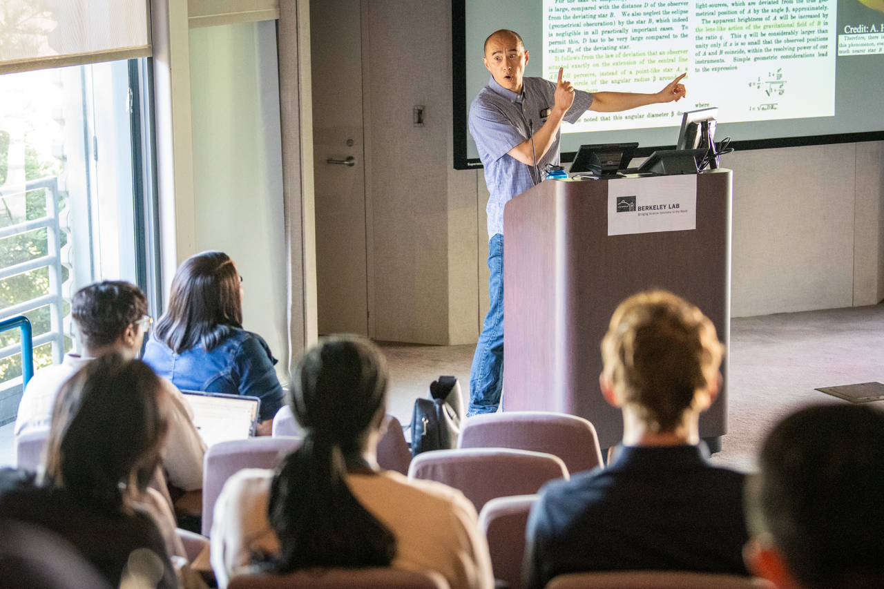 Person lecturing from podium gesturing toward projected image on wall with seated people listening.