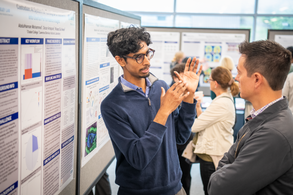 Person standing in front of poster board gesturing to another person while engaged in conversation with other posters and people in the background.