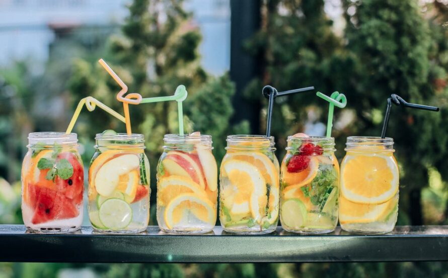 Mason jars in a line on top of a table containing brightly colored fruit slices and liquid with curly straws