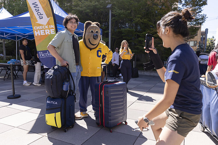 UC Berkeley student smiling and posing with Cal Bear mascot while holding suitcase.