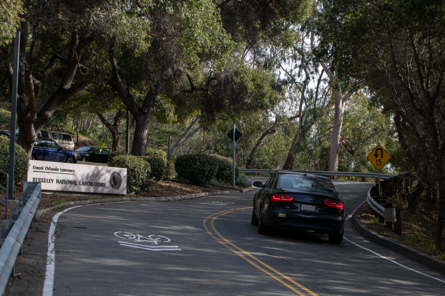 Winding road with a black sedan driving up next to a sign for Lawrence Berkeley National Laboratory surrounded by trees and foliage.