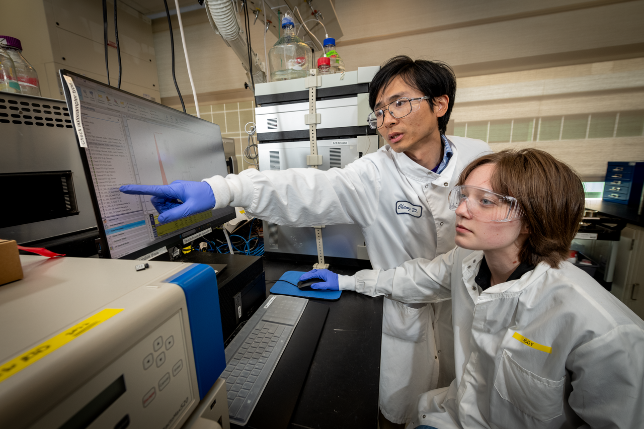 Two science researchers wearing lab coats and protective equipment look at computer monitor as one researcher gestures toward the screen.