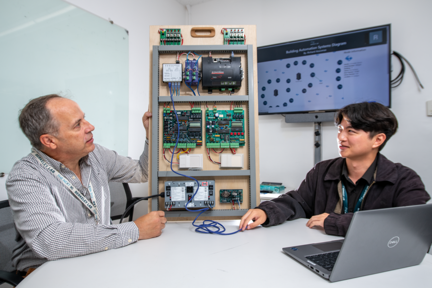 Two persons seated at table in front of laptop computer engaged in discussion with scientific instruments in the background.