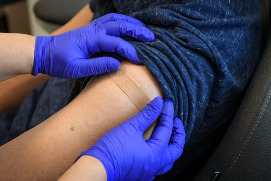 Nurse with gloved hands applying an adhesive bandage to a patient’s arm post-vaccination.