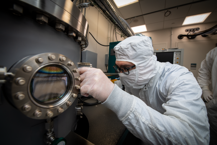 Person in white full-body hazard suit wearing black glasses adjusting scientific instrumentation.