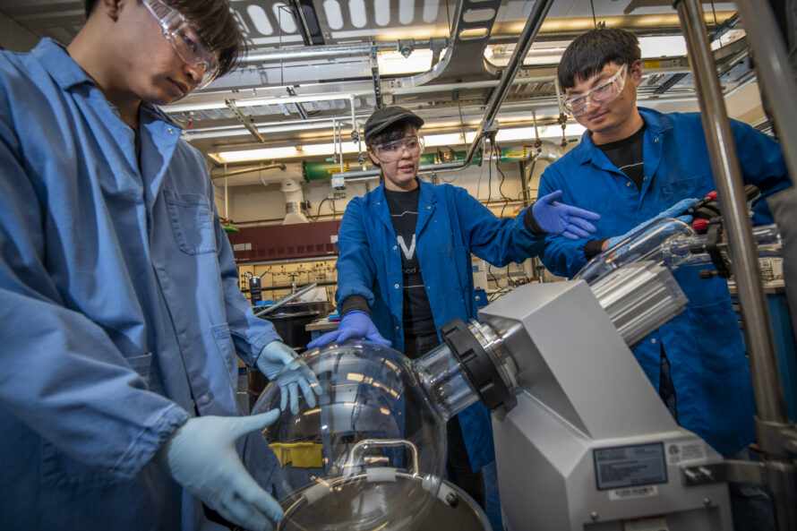 Three scientists in blue lab coats and goggles huddle around a piece of scientific equipment in a laboratory, with one of them handling a glass apparatus connected to the equipment.