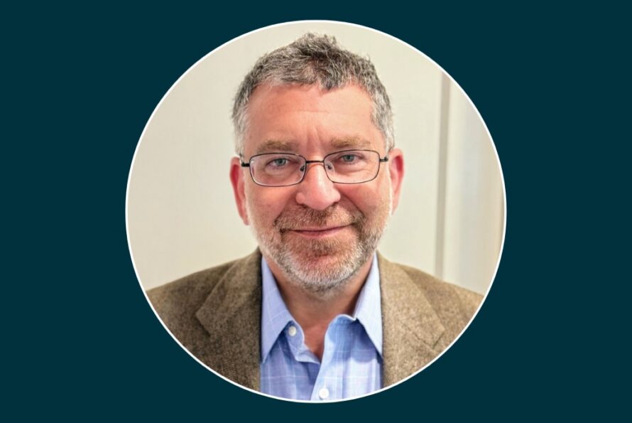 Portrait of Adam Stone, a person with gray hair and beard wearing glasses and a beige jacket over a blue collared shit.