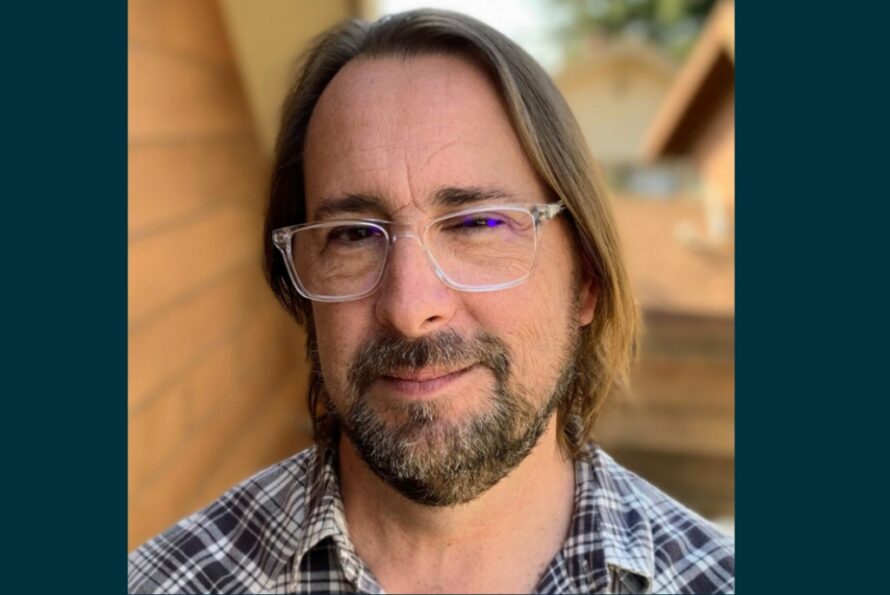 Headshot of person with long brown hair, wearing glasses and a black and white plaid shirt.