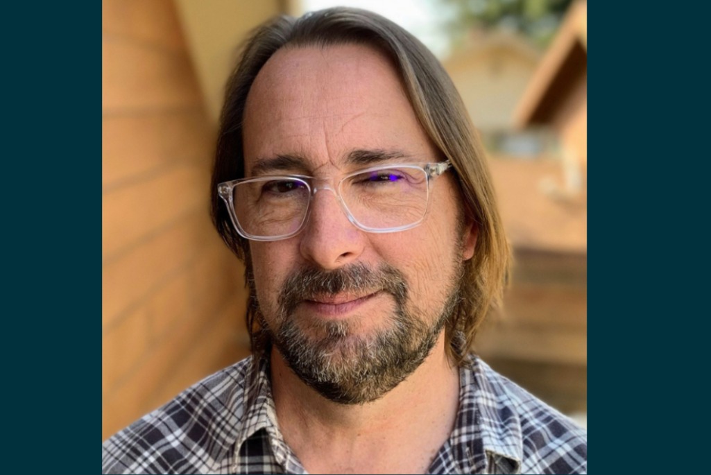 Headshot of person with long brown hair, wearing glasses and a black and white plaid shirt.