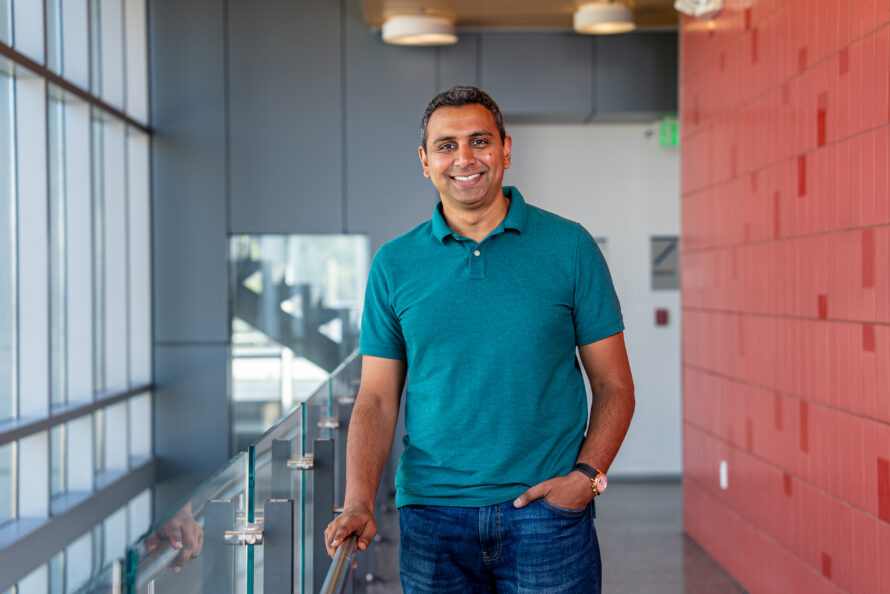 Researcher at Berkeley Lab with short dark hair wearing blue shirt stands between sunlit windows and a red wall.
