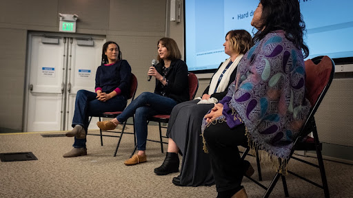 Four persons seated as panelists in front of large screen speaking to audience.
