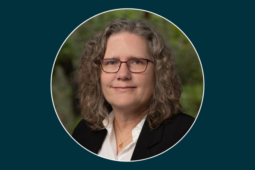 Portrait of Lori Zscherpel, a person with curly brown hair wearing glasses, necklace, and a black blazer over a white collared shirt.