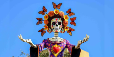 Multi-colored Día de los Muertos Catrina skeleton figure against blue sky.