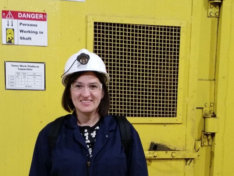 Engineer with shoulder-length brown hair and black sweater wearing protective lab hat and eyeglasses.