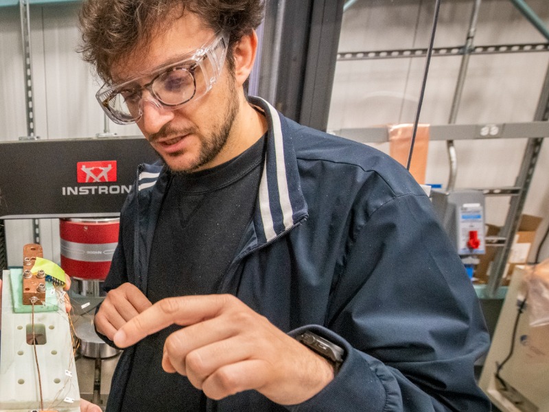 Research scientist with brown hair and beard wearing dark blue t-shirt and jacket adjusting a device in a lab setting.